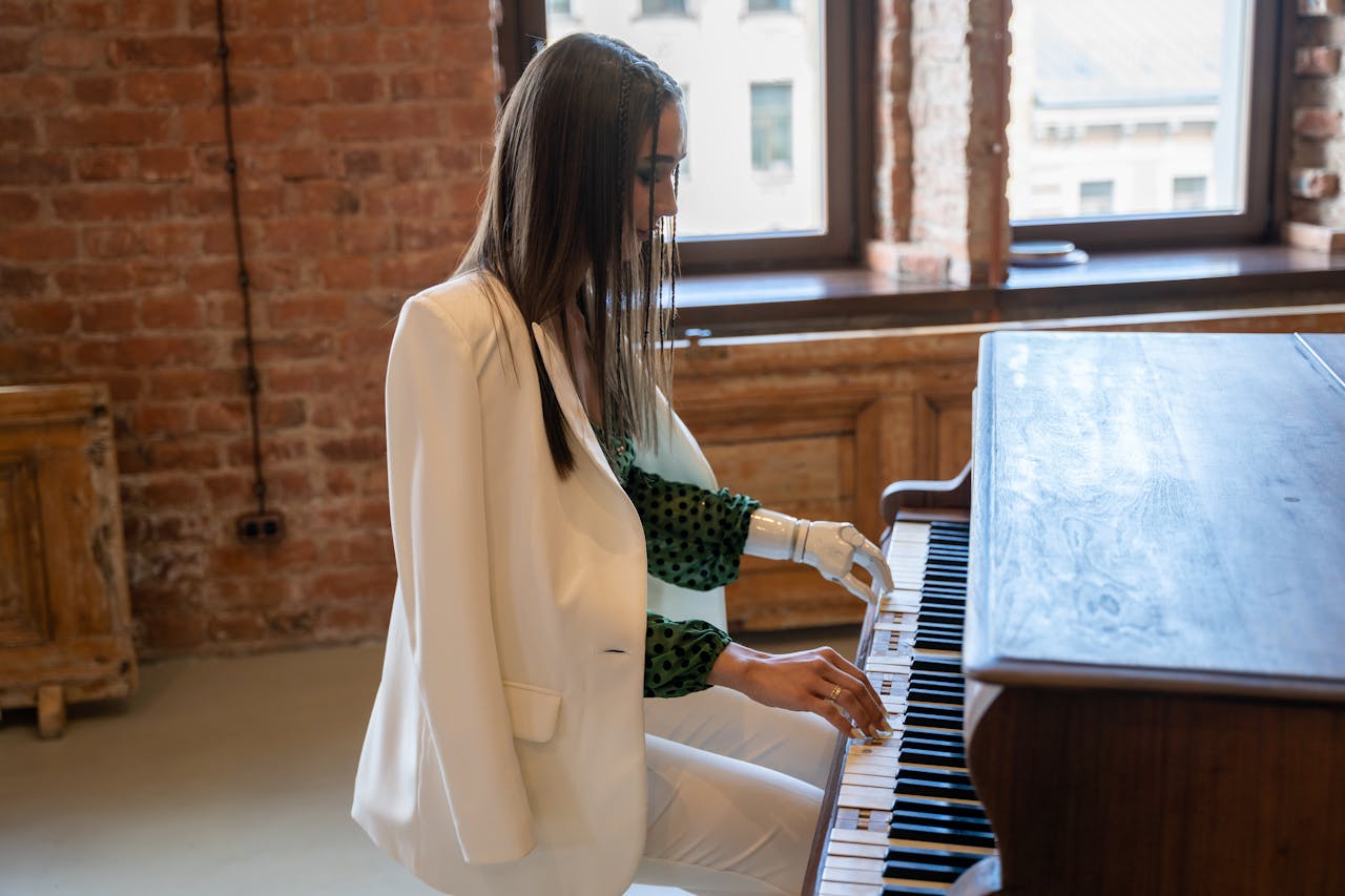 A woman in a white blazer with a prosthetic hand playing piano in a brick-walled room.